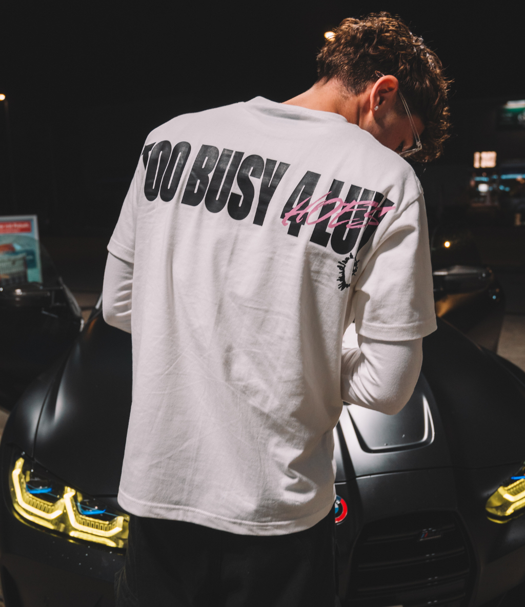 Model wearing NOLUV TEE white cotton t-shirt with "TOO BUSY 4 LUV" print, standing in front of a stylish car.