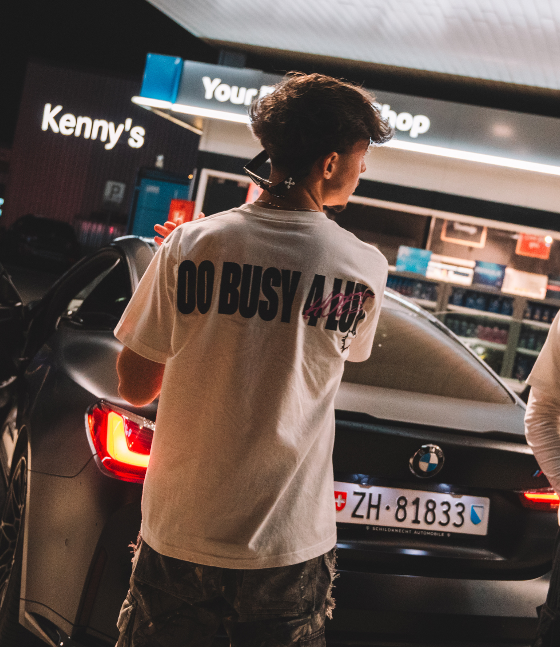 Young man wearing a white cotton NOLUV tee with "Too Busy 4 Luv" print, standing near a BMW at night.
