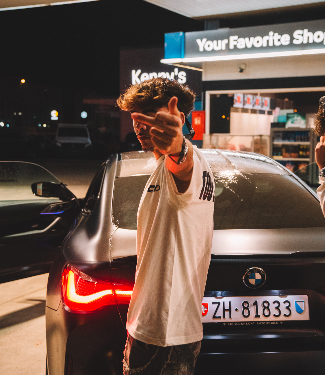 Young man wearing a white cotton T-shirt with a unique design, posing next to a sleek black car at night.
