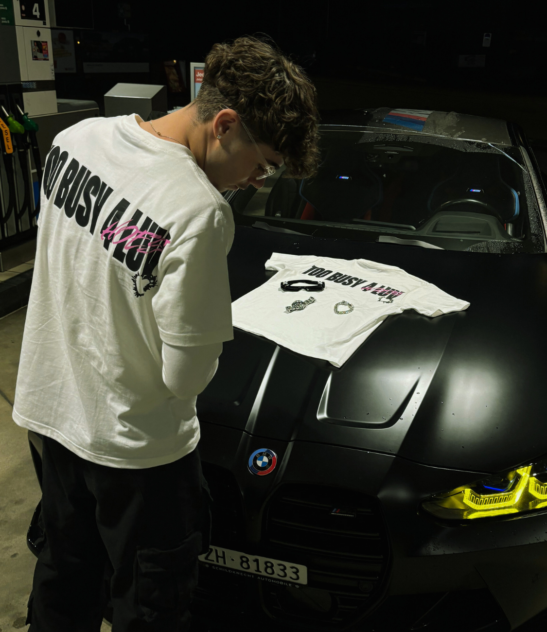 Unisex white NOLUV tee with "Too Busy 4 Luv" design displayed on a BMW at night, featuring Chasedvision keychain and stickers.