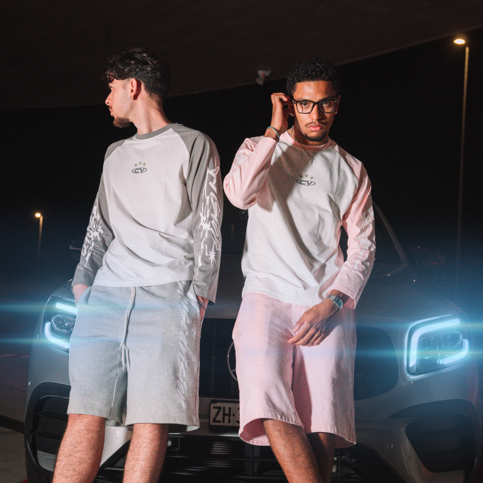 Two men wearing summer longsleeve shirts and pink shorts, posing by an SUV at night.