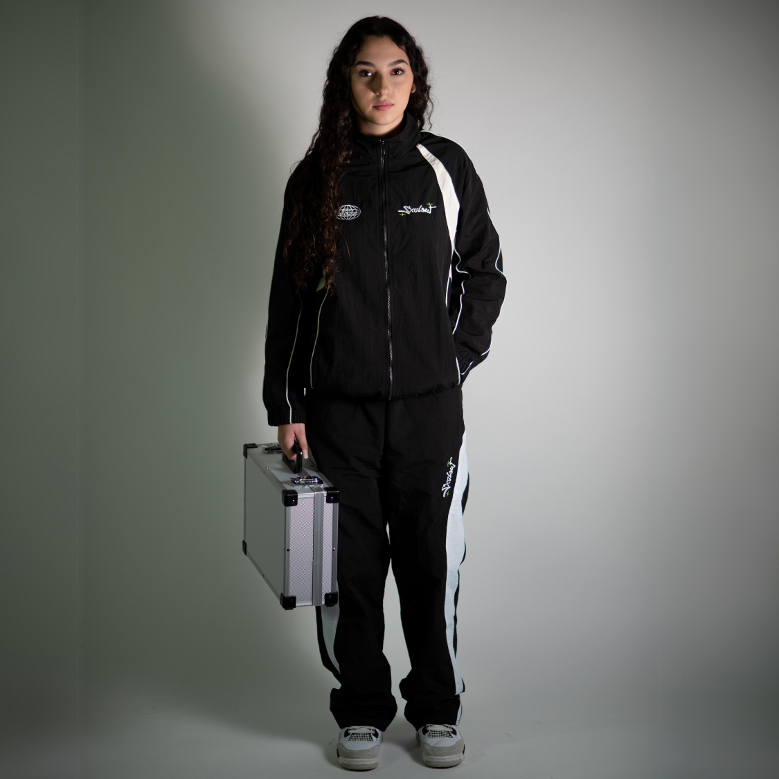 Person wearing black chasedvision nylon tracksuit bundle with trackjacket and trackpants, holding a silver case, stylish streetwear fashion.