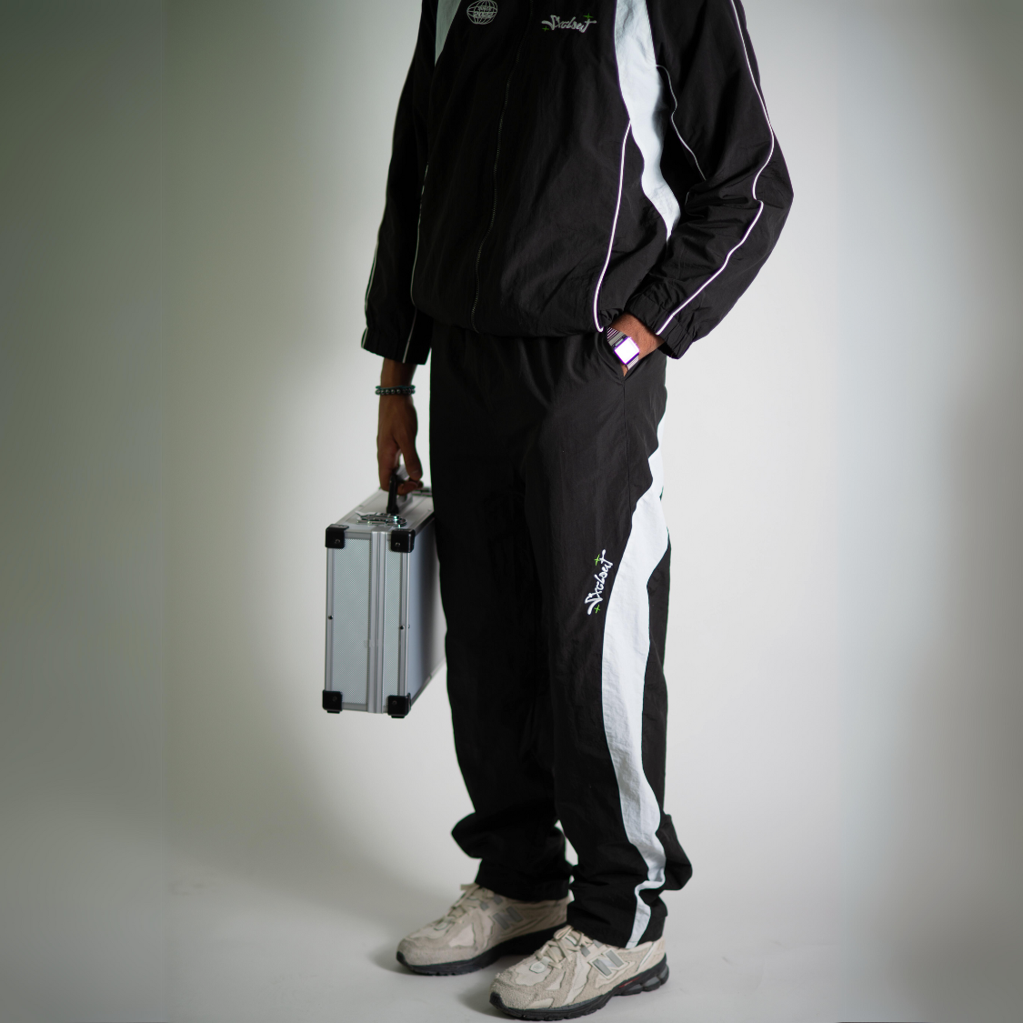 Person wearing a black nylon tracksuit bundle with trackjacket and trackpants, holding a silver briefcase; stylish streetwear by chasedvision.