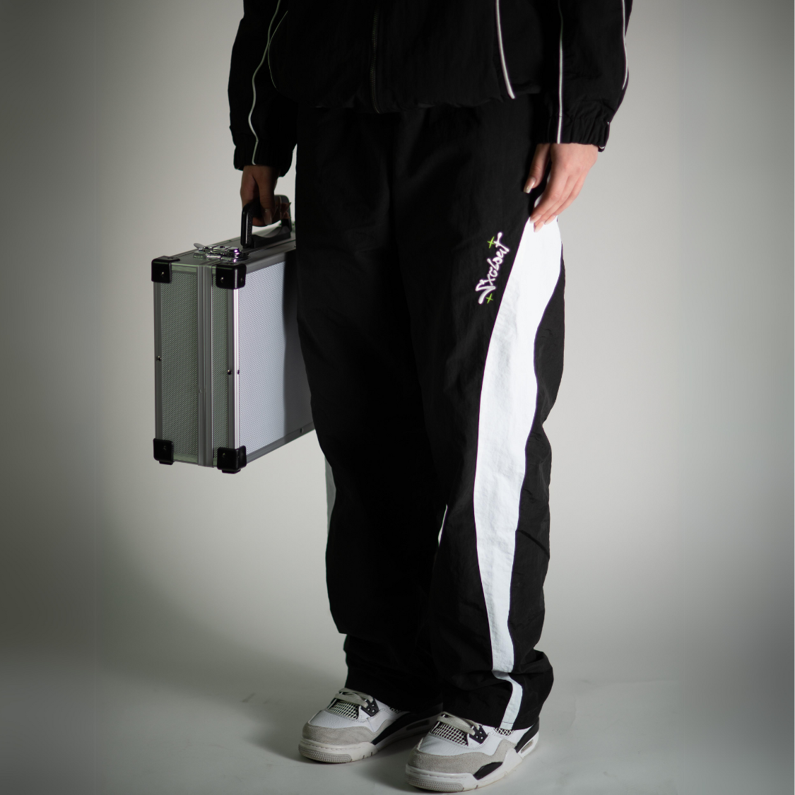 Person wearing Vision Trackpants, black baggy nylon streetwear by Chasedvision, holding a silver briefcase.