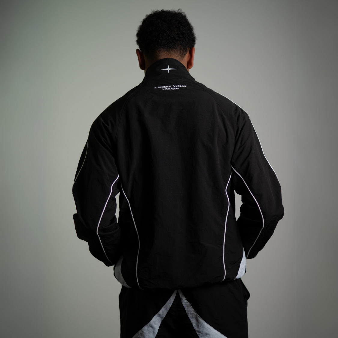 Back view of a man wearing black VISION TRACKJACKET, nylon streetwear, designed by Chasedvision, showcasing reflective accents.