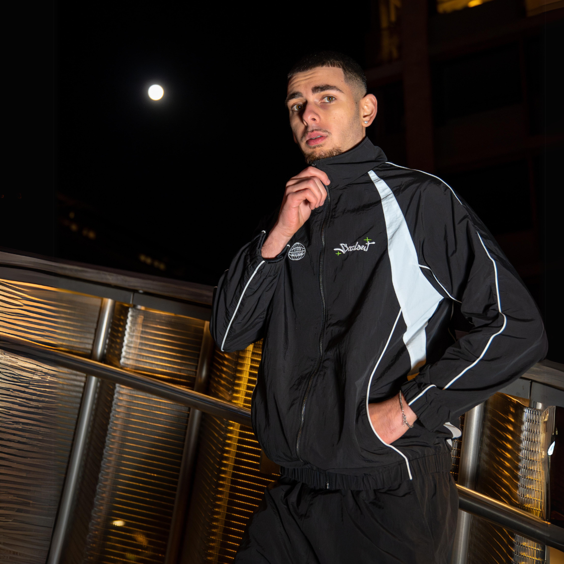 Man wearing VISION TRACKJACKET, a black nylon streetwear jacket by Chasedvision, standing outdoors at night under a full moon.