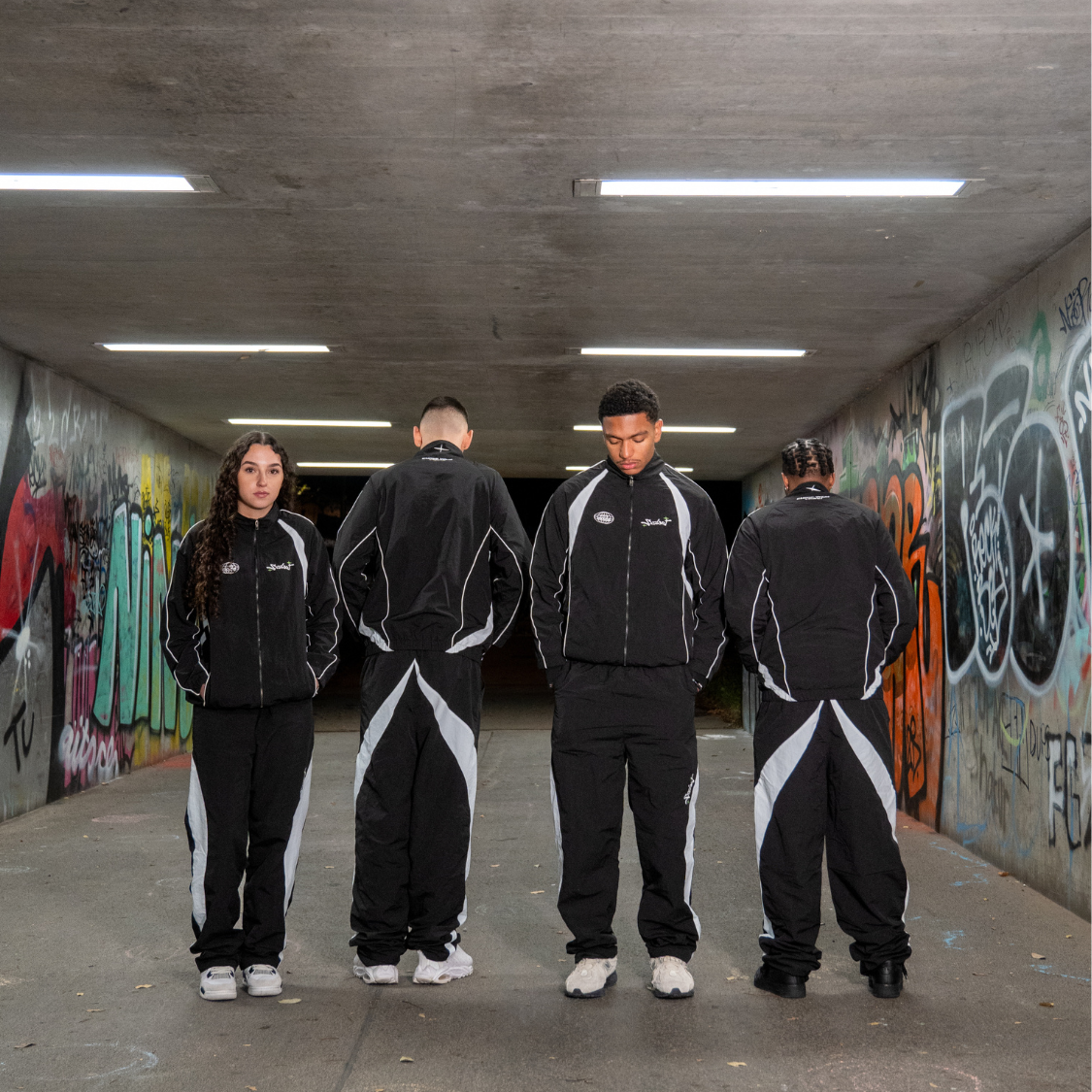 Group wearing Chasedvision nylon baggy streetwear trackpants in graffiti-covered urban setting.