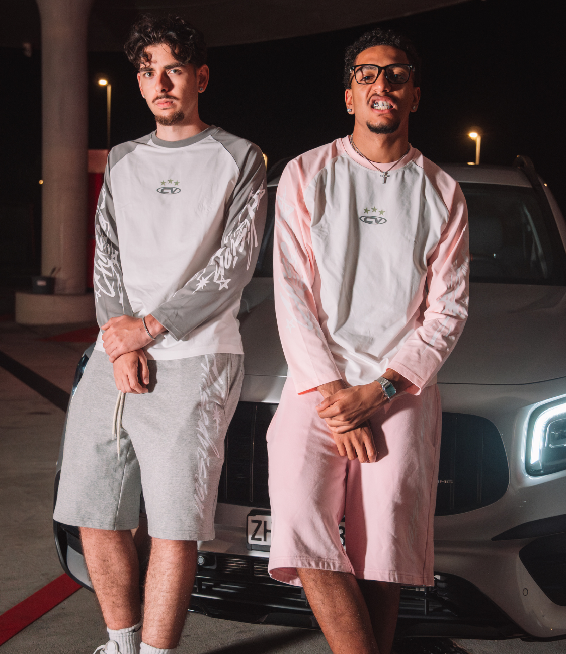Two men wearing summer longsleeve shirts and pink shorts, posing by an SUV at night.